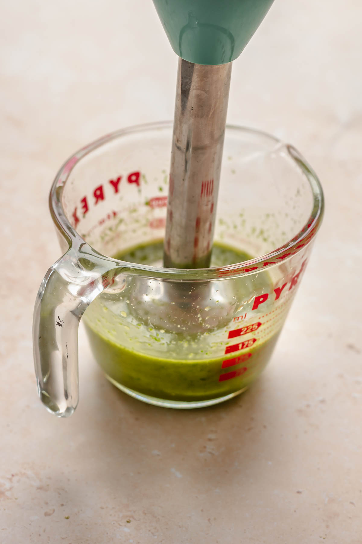 An immersion blender blends basil and garlic oil in a glass measuring cup.