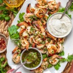 A hand spoons chimichurri on a platter of grilled shrimp.