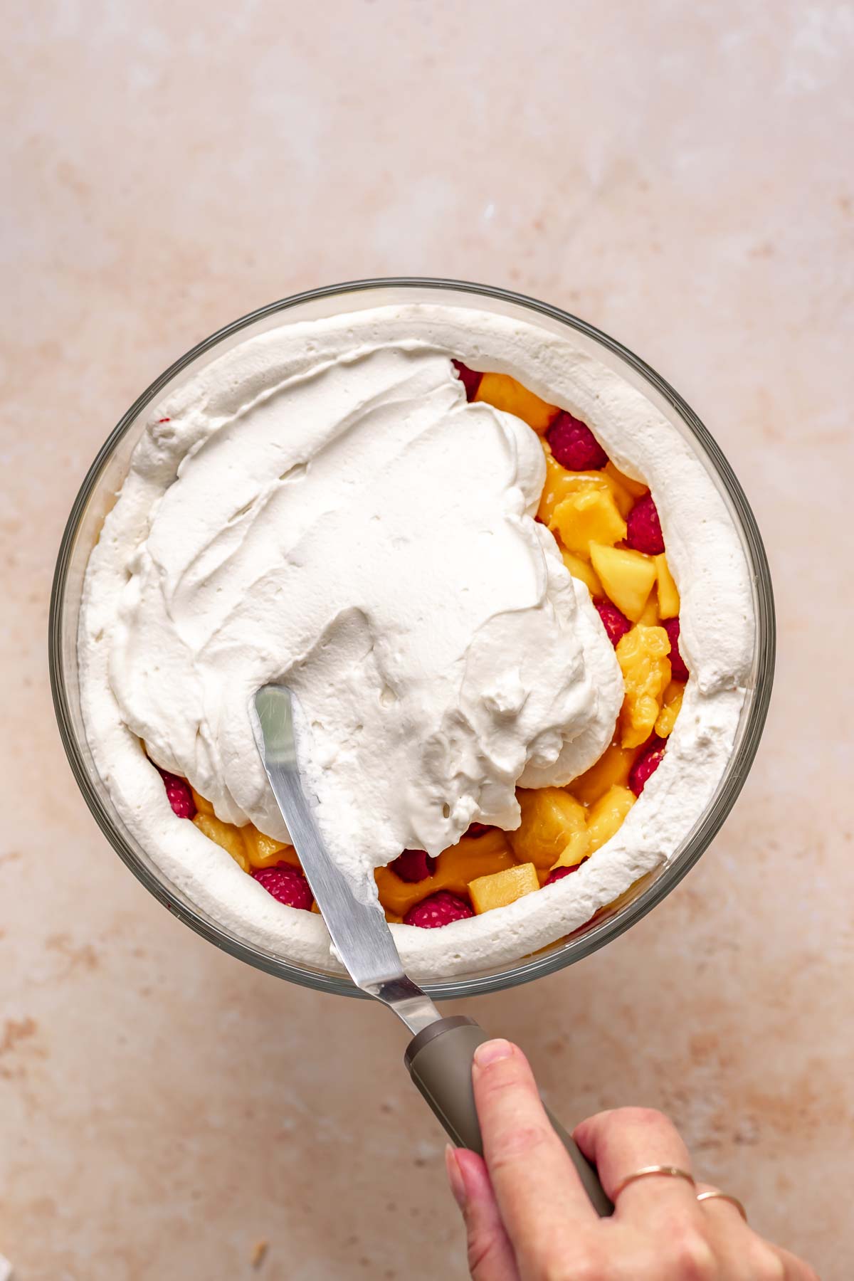 Coconut whipped cream being spread on top of fruit in the bowl for the final layer.