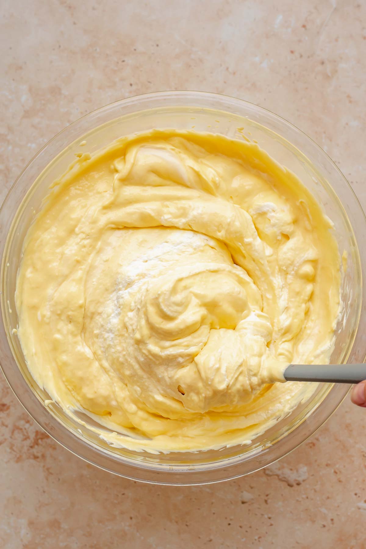 A spatula folds whipped cream into mango cheesecake batter.