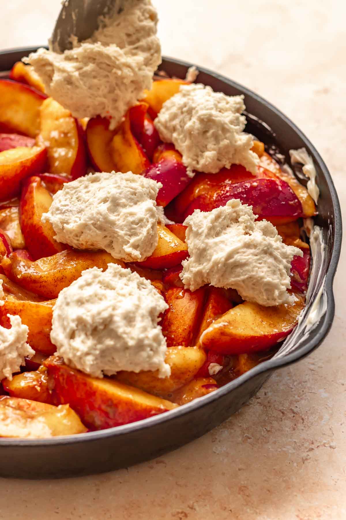 A spoon adds dollops of biscuit dough on top of sliced peaches.