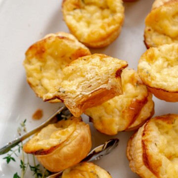 Tongs hold a cheese tartlet on a platter with a bite removed.