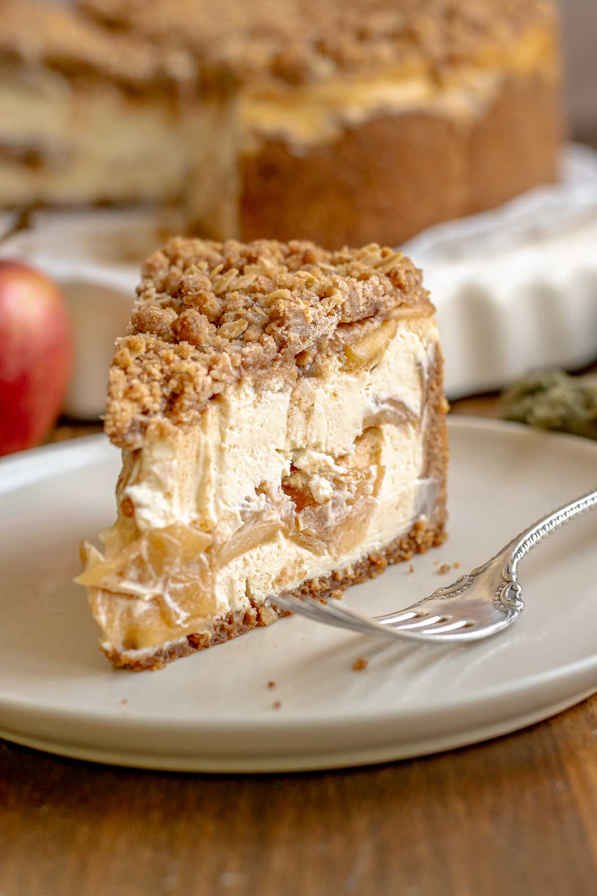A slice of apple crisp cheesecake on a plate with a fork next to it.