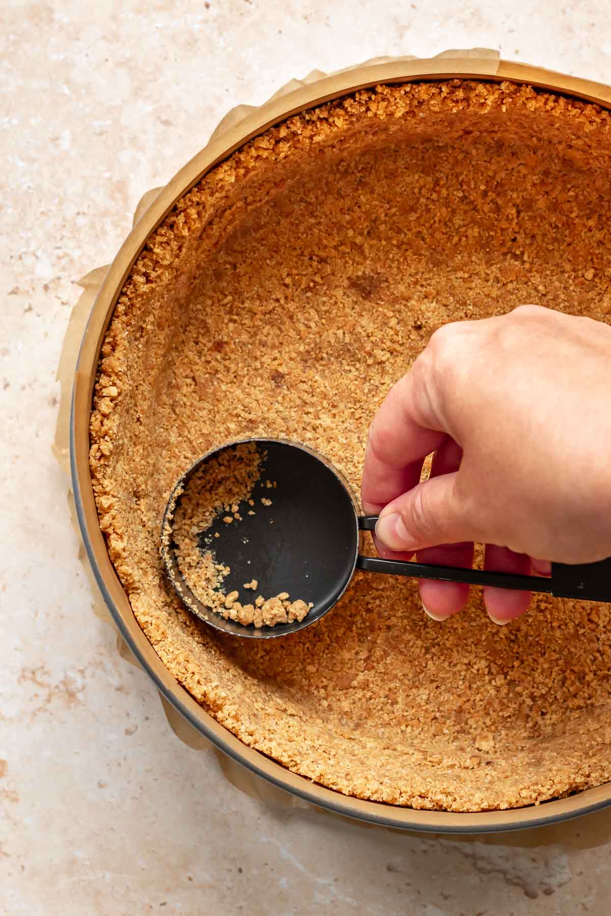 A hand uses a measuring cup to press graham cracker crumbs into a springform pan.