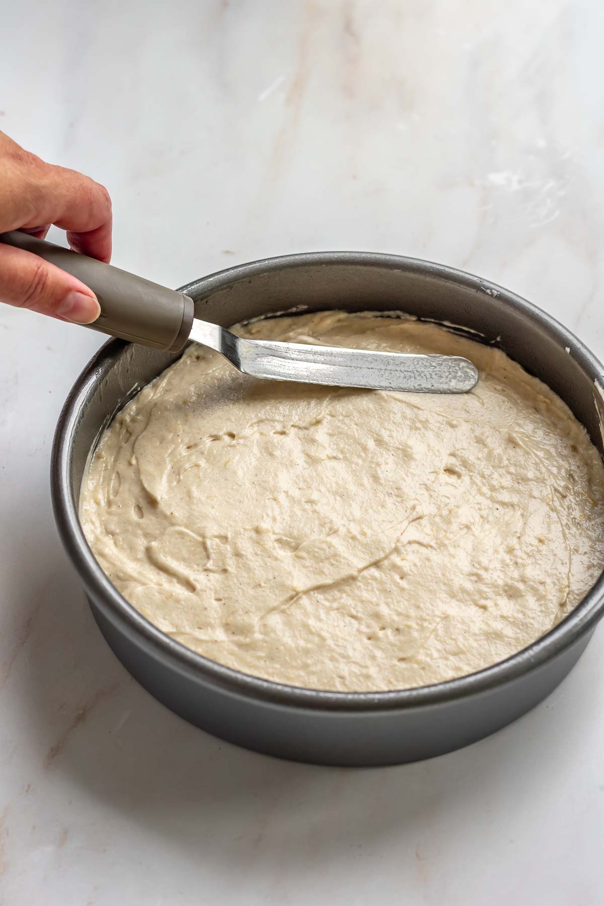 A hand uses an offset spatula to spread cake batter into a cake pan.