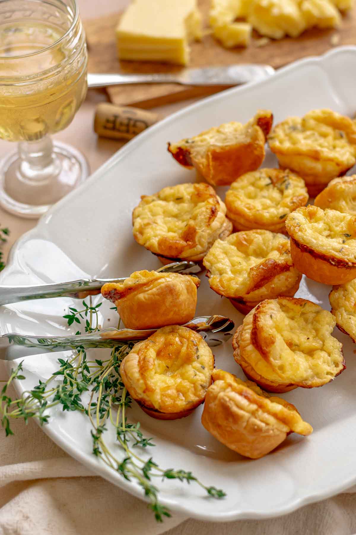 Tongs reach in to a platter of cheese tartlets. Wine and thyme sit on the site.