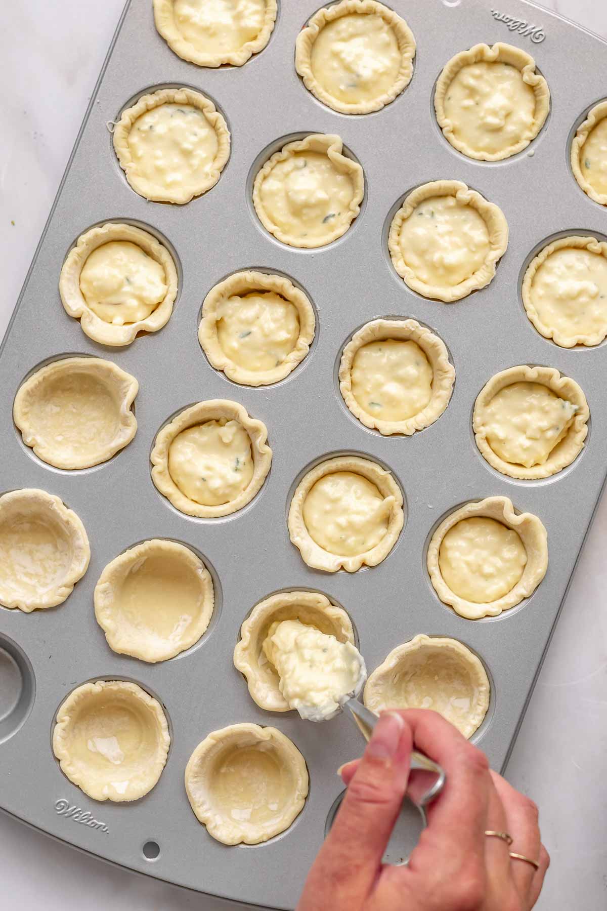 A spoon adds cheese filling into puff pastry shells in a mini muffin tin.