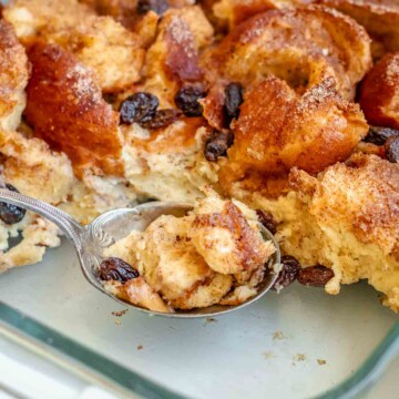 A serving spoon holds a spoonful of cinnamon bread pudding in a casserole dish.
