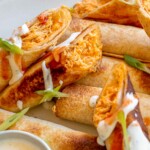 Buffalo chicken taquitos on a platter drizzled with ranch. Some are cut open.