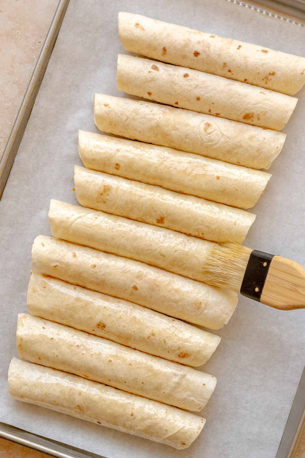 A brush spreads olive oil on top of rolled buffalo chicken taquitos.