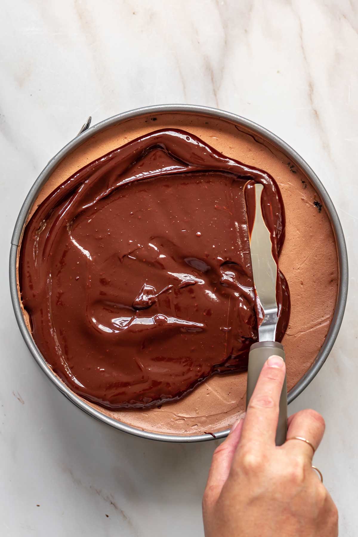 An offset spatula spreads chocolate ganache on top of cheesecake.