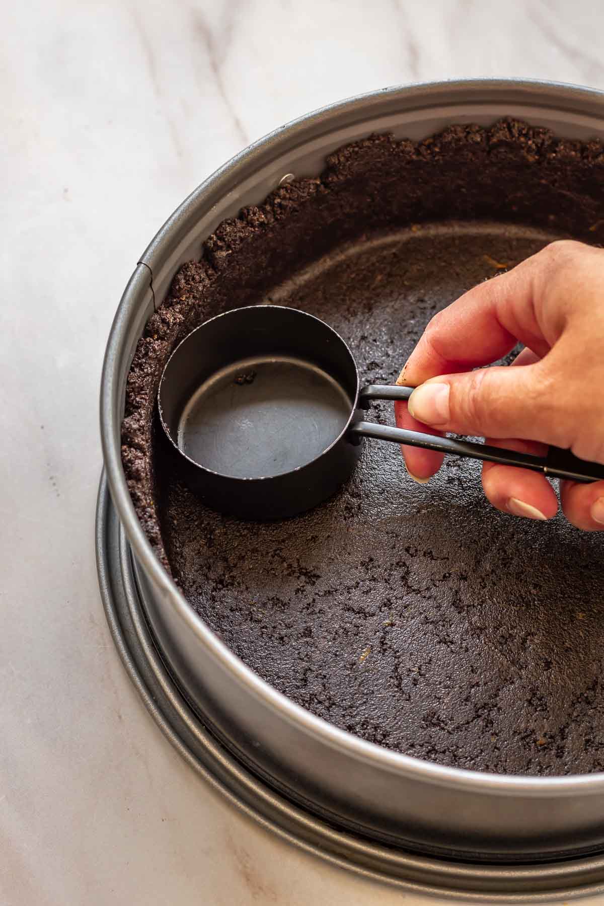 A hand uses a measuring cup to press oreo crumbs into a springform pan.