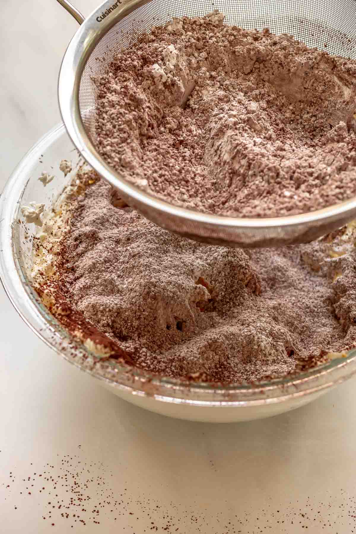 Cocoa powder and sugar being sifted into a bowl of cream cheese.