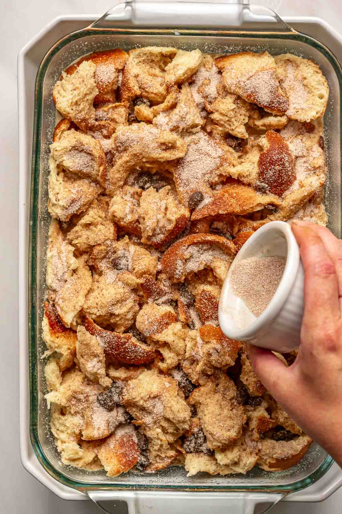 A hand sprinkles cinnamon sugar on top of the bread pudding before baking.