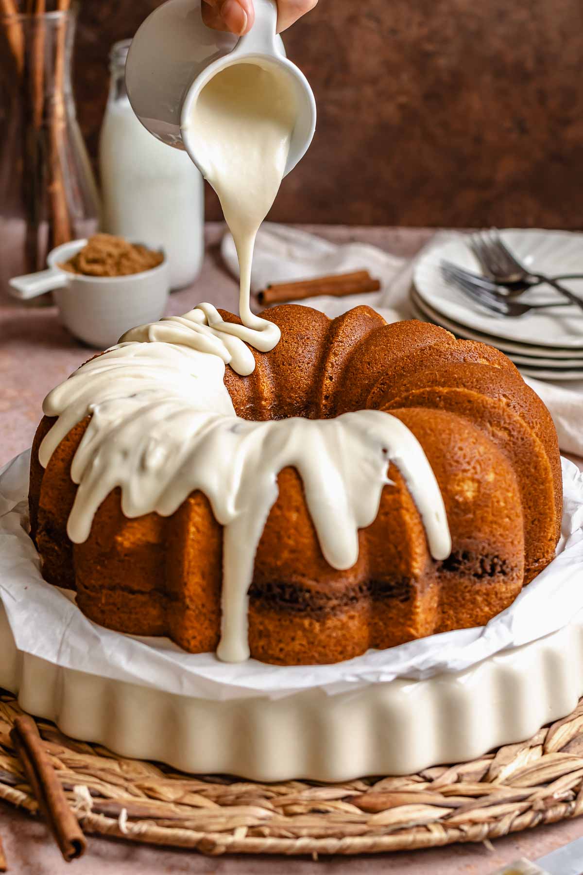 A hand pours cream cheese frosting over a cinnamon bundt cake on a platter.
