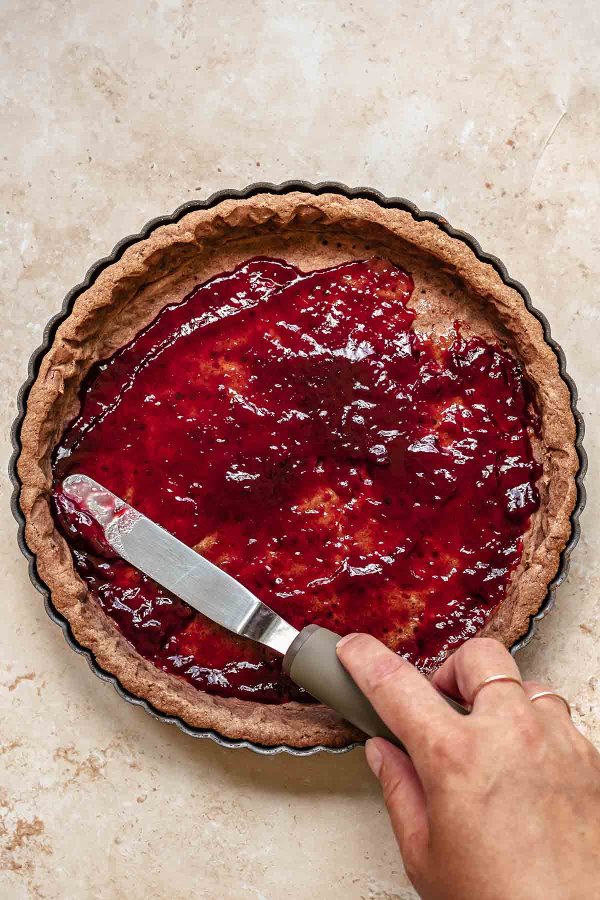 An offset spatula spreads raspberry jam onto a cooked tart crust.