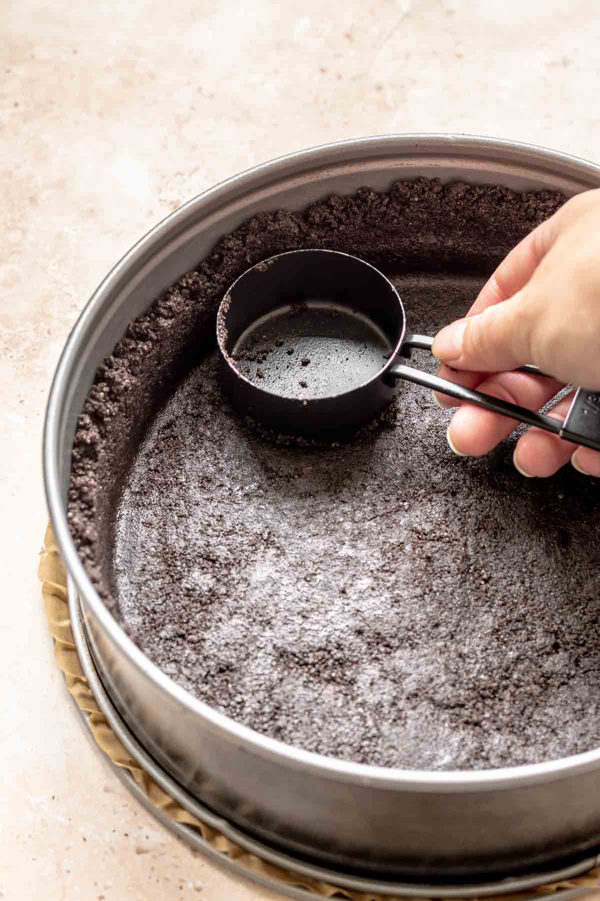 A hand presses oreo crumbs into a springform pan.