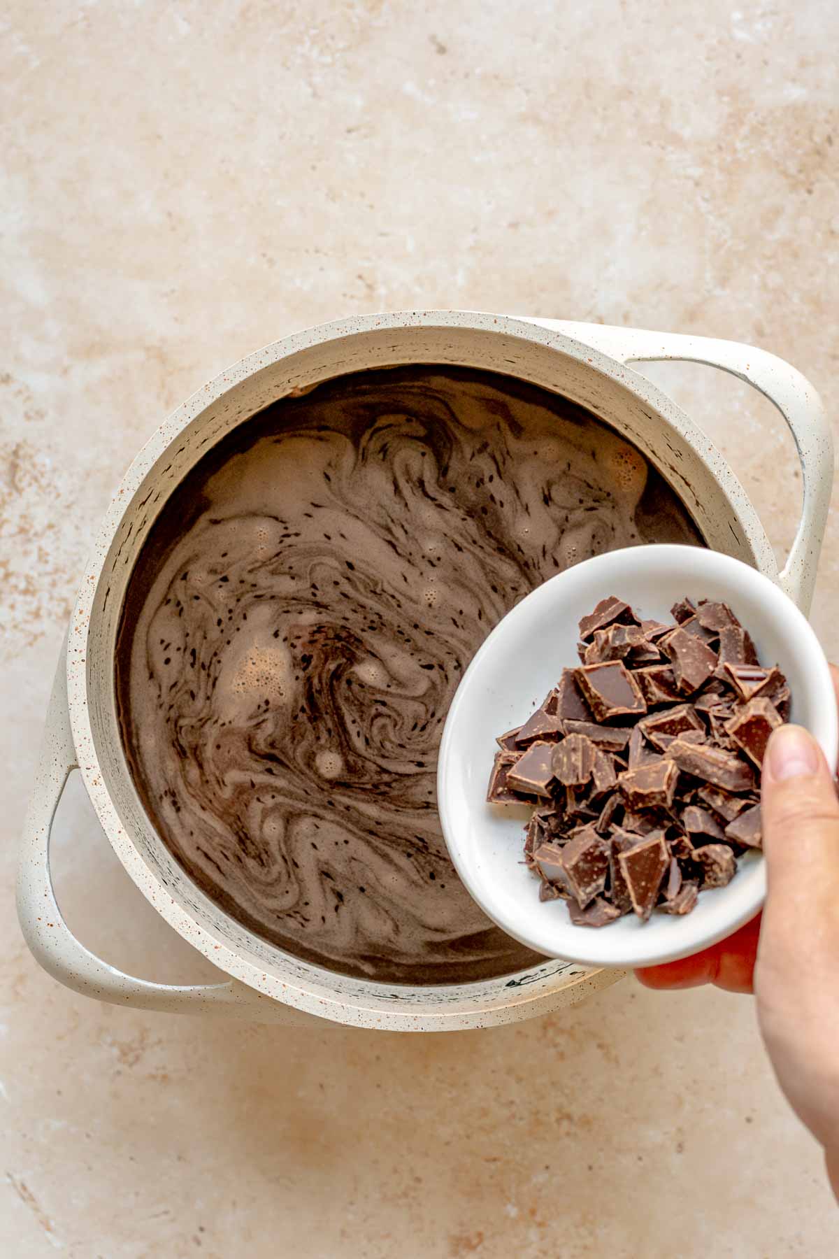 A hand pours chopped chocolate into hot cocoa in a saucepan.
