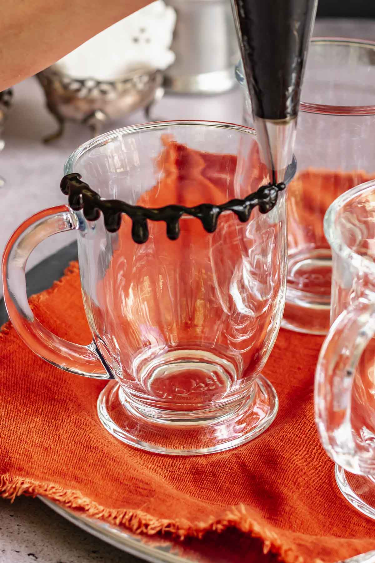 A piping tip adds drips of black hot fudge onto the rim of a clear mug.