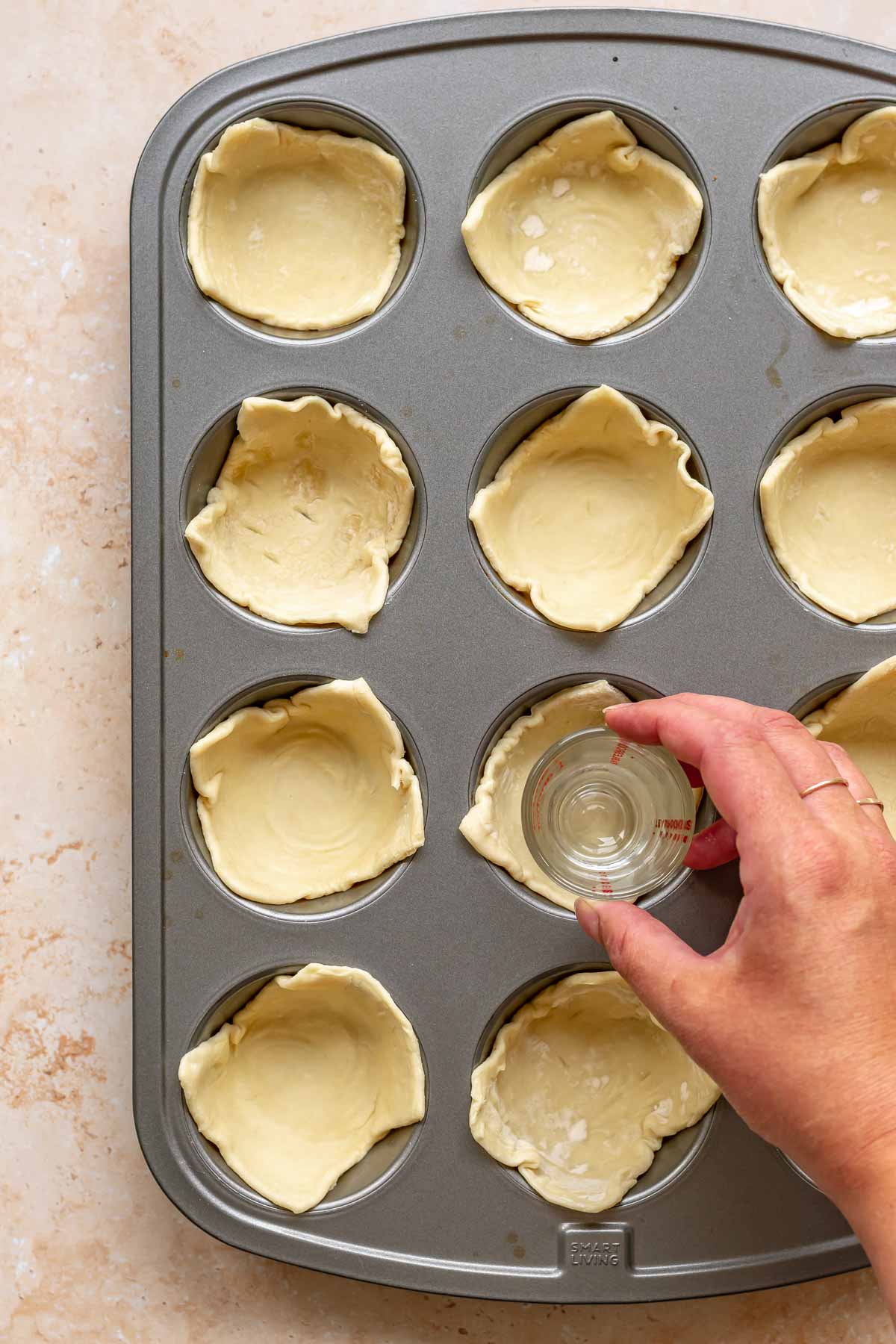 A hand uses a shot glass to press squares of puff pastry into a muffin tin.