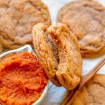 Pumpkin cookies leaning on a cup of pumpkin puree. One has a bite removed.