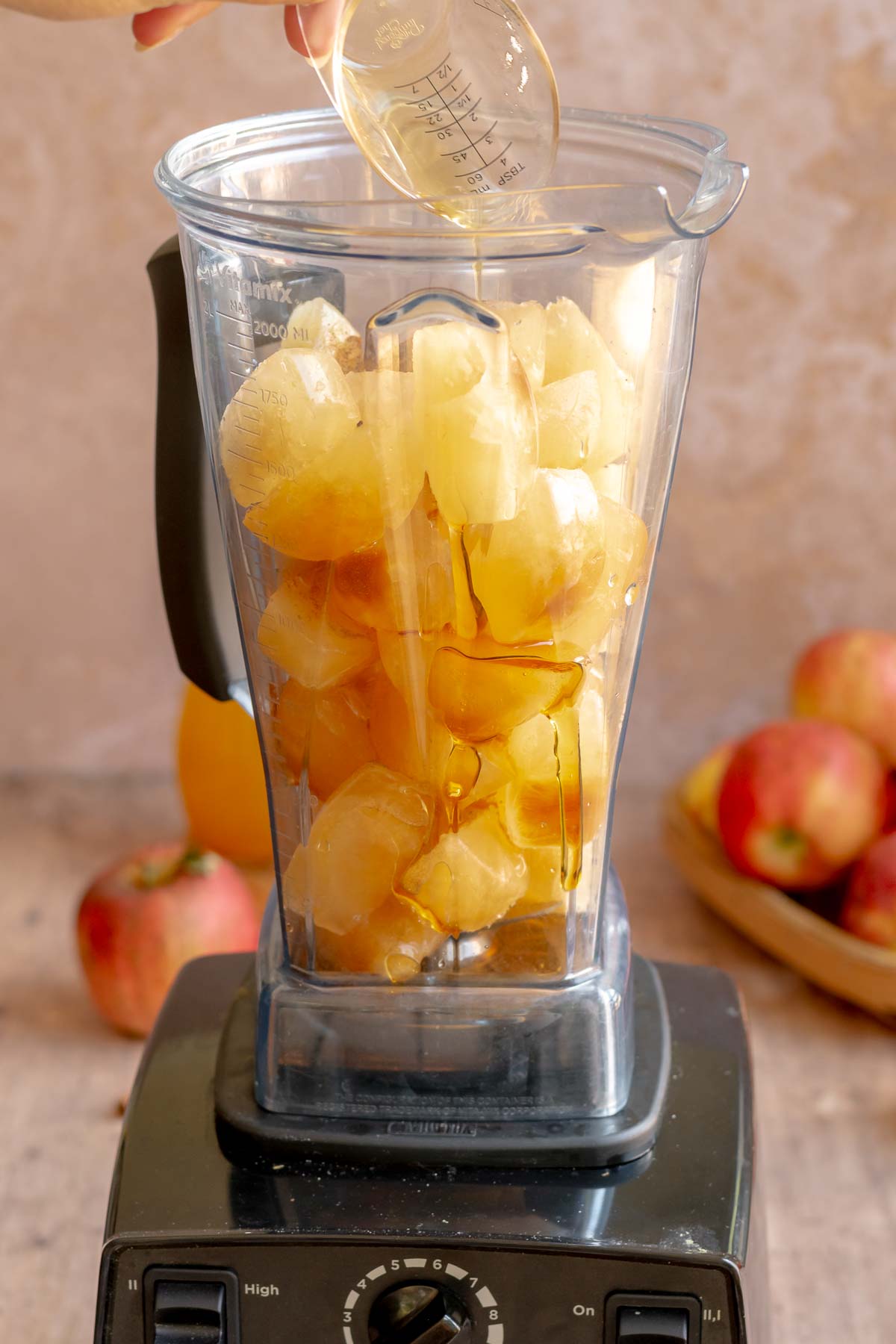 A hand pours maple syrup into a blender full of apple cider ice cubes.