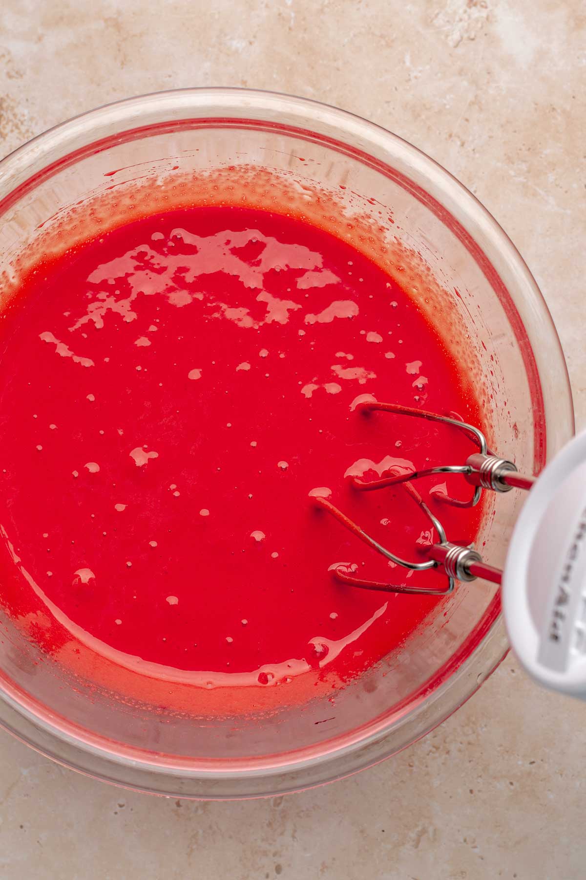 A hand mixer mixes red batter in a bowl.