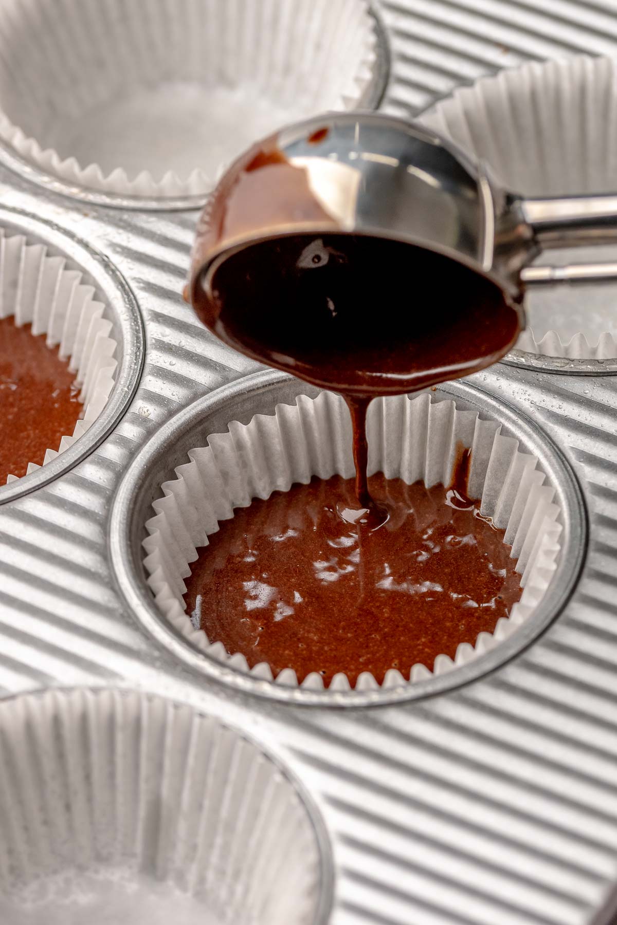 A cookie scoop adds chocolate cake batter into a muffin tin.