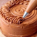 A piping bag pipes chocolate swiss meringue buttercream onto a cake.