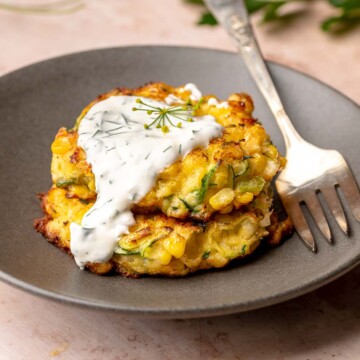 A stack of two corn and zucchini fritters on a plate with yogurt sauce dripping down the side.