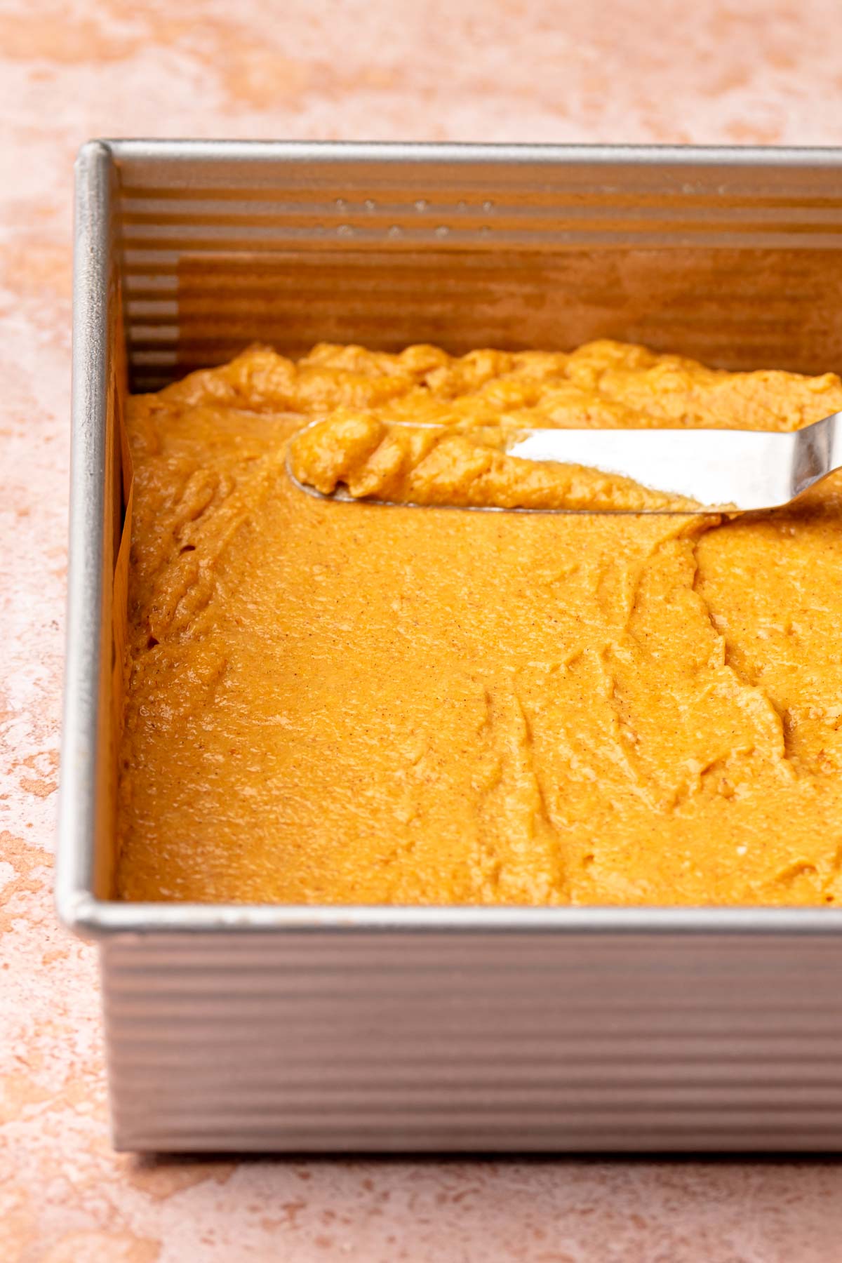 A spatula spread pumpkin cake batter into a prepared square pan.