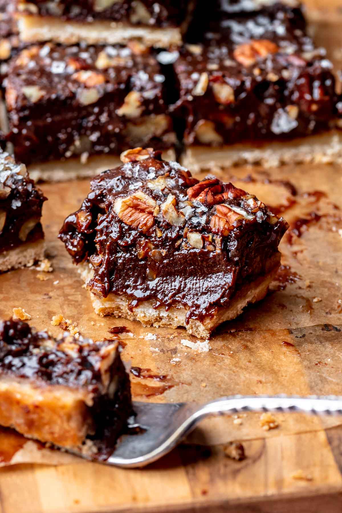A slice of chocolate pecan pie bar on a piece of parchment paper. A bite is removed on a fork.