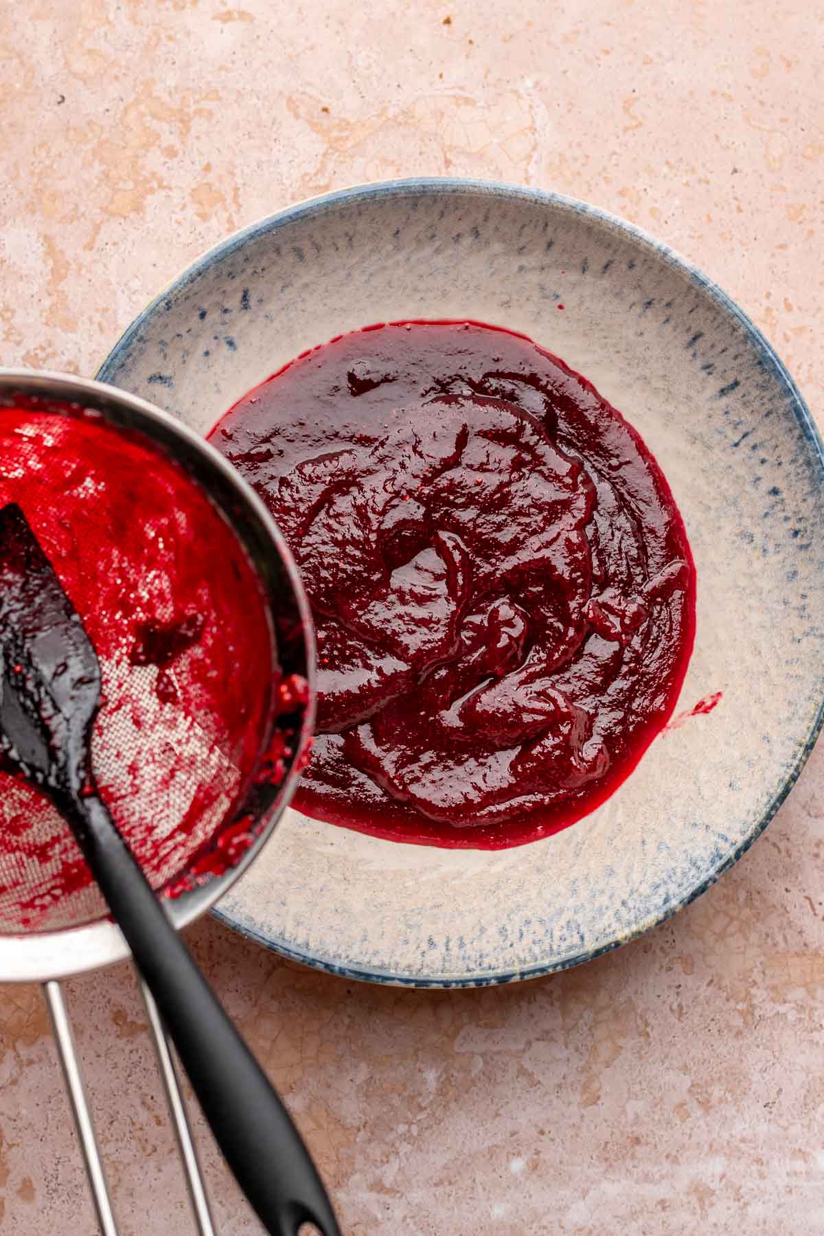 A spatula pushes cranberry puree through a fine mesh sieve into a bowl.