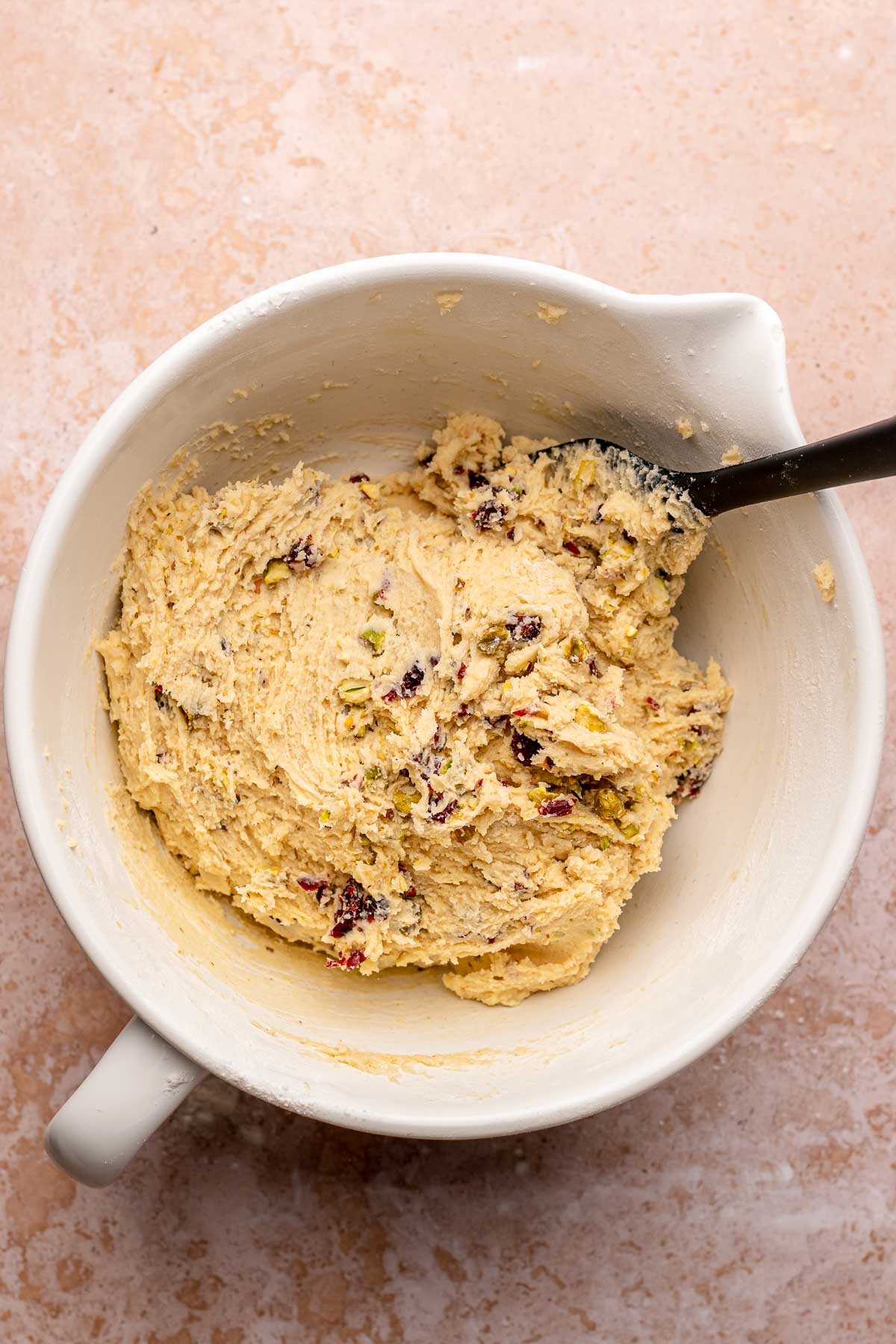 A spatula mixes cranberry pistachio dough in a bowl.