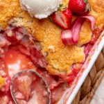 A spoon sits in a casserole dish of strawberry rhubarb cobbler with some removed.
