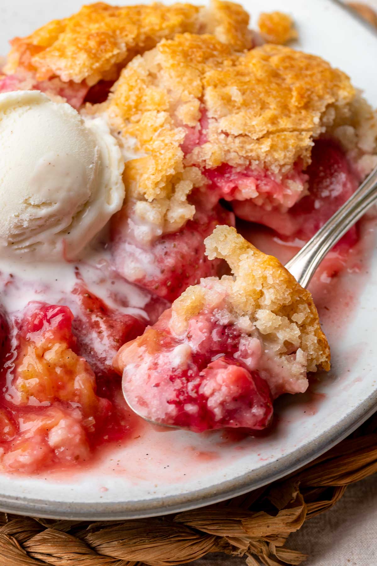 Strawberry rhubarb cobbler on a plate with a scoop of vanilla ice cream on top. A spoon holds a scoop.