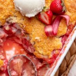 A spoon sits in a casserole dish of strawberry rhubarb cobbler with some removed.