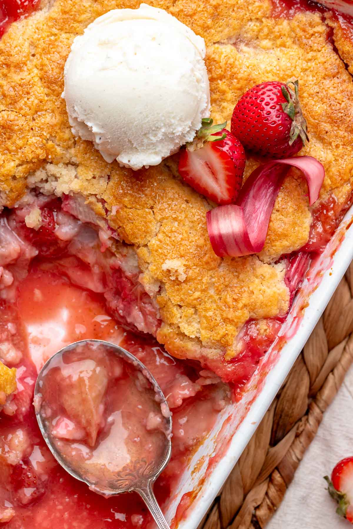 A spoon sits in a casserole dish of strawberry rhubarb cobbler with some removed.
