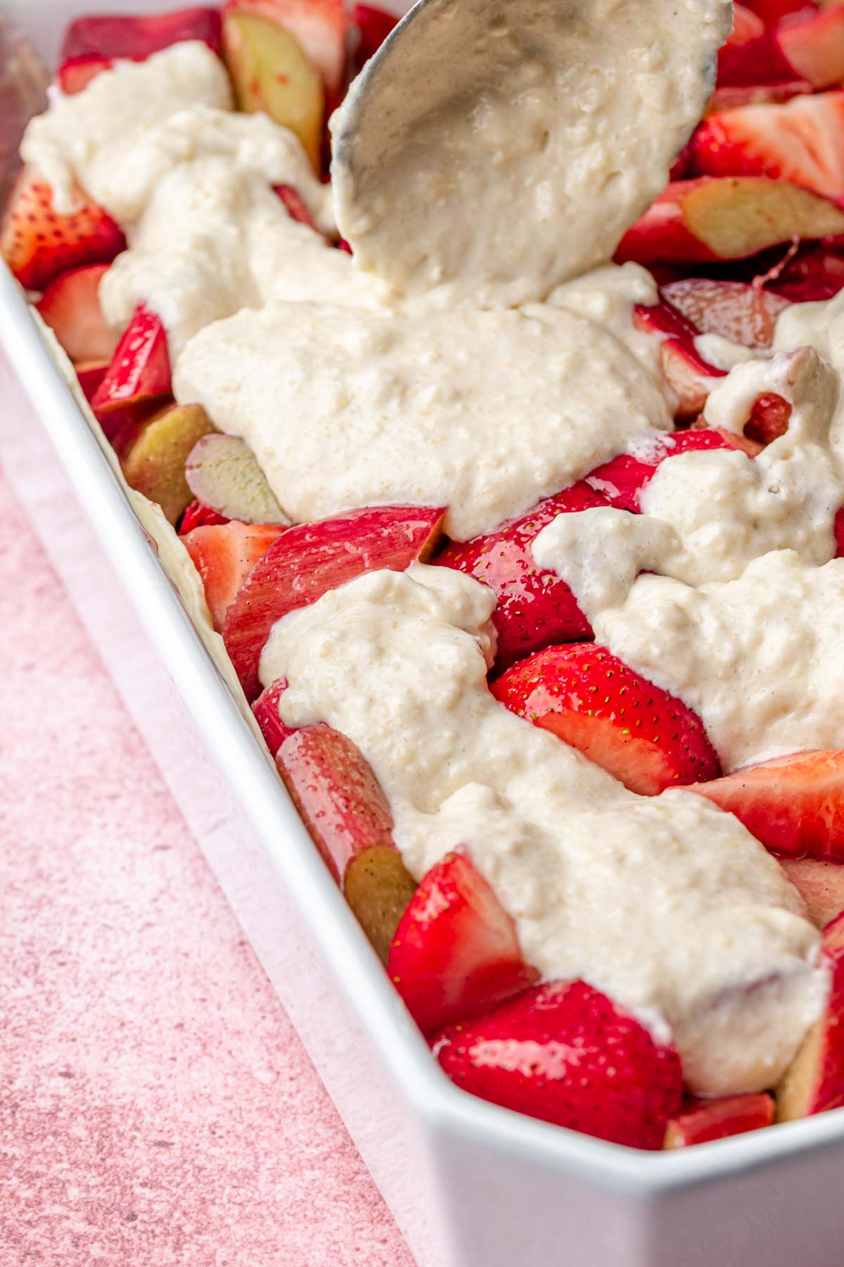 A spoon pours batter on top of strawberries and rhubarb in a casserole dish.