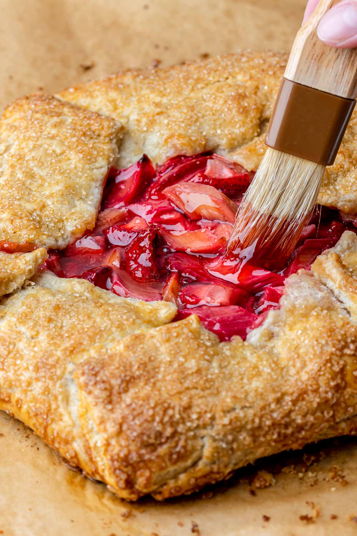 A pastry brush adds strawberry jam onto the fruit of a baked galette.