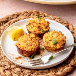 Three mini crab cakes on a plate with a fork and lemon wedge.