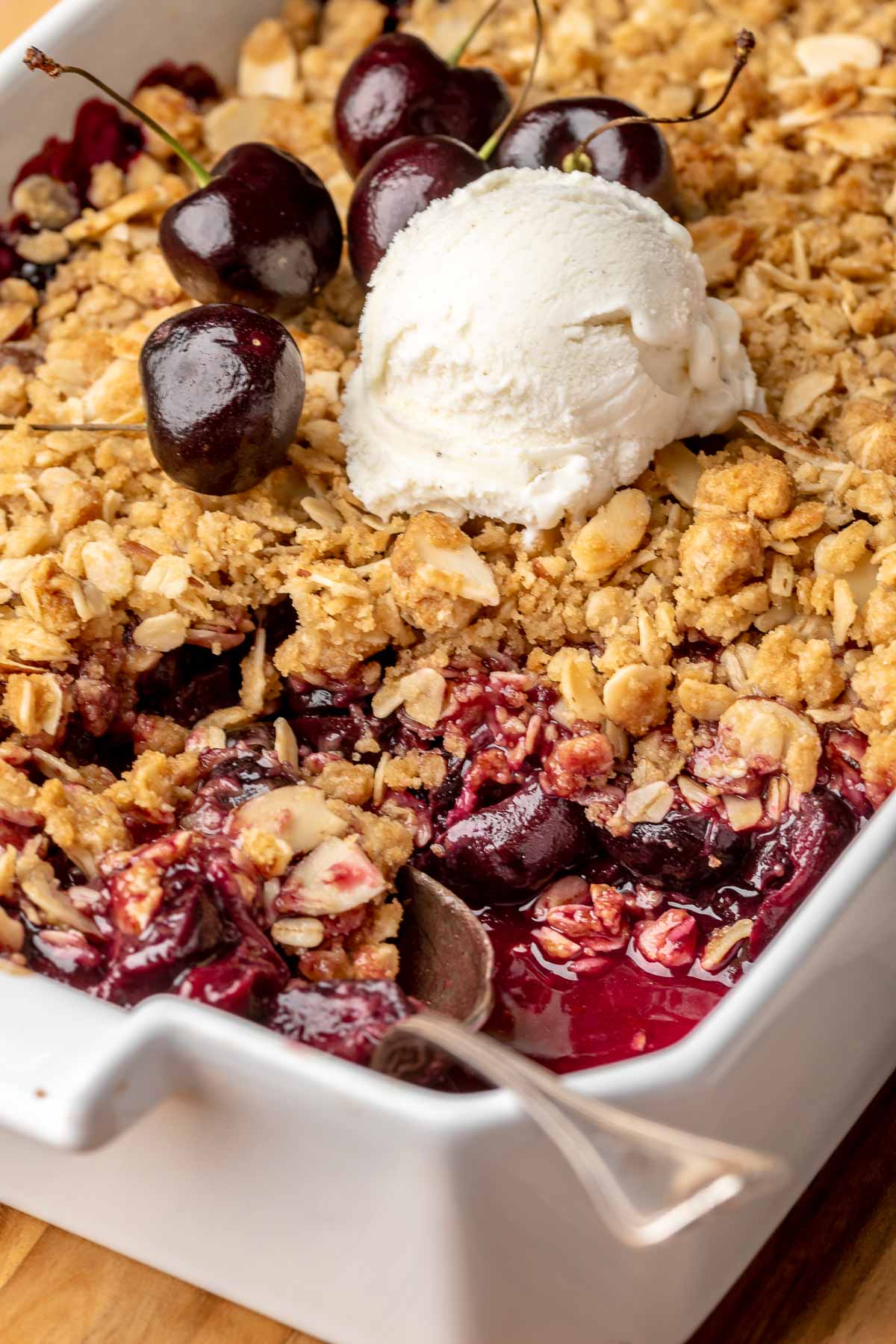 Cherry crisp in a casserole dish with a spoonful removed and a scoop of ice cream on top. A spoon sits in the corner.