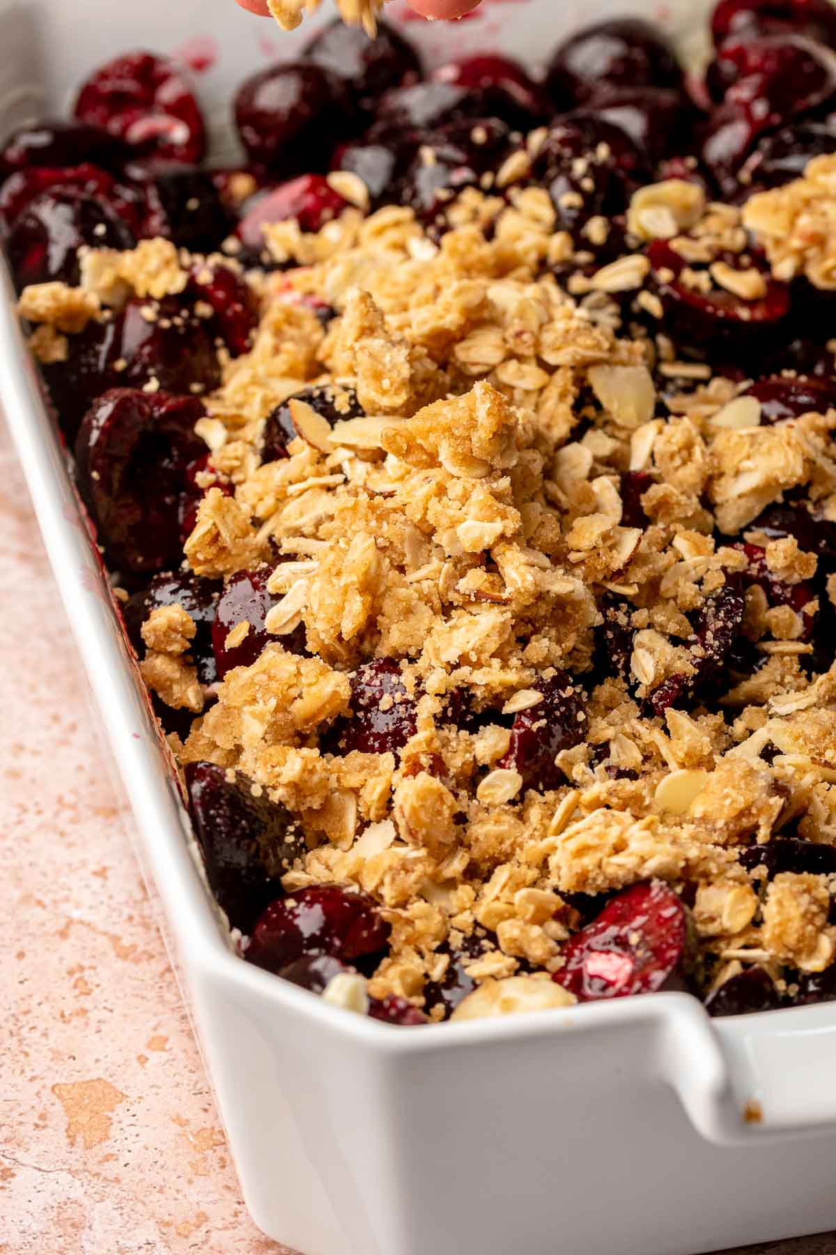 A hand sprinkles oat crumbles on top of the cherries in a casserole dish.