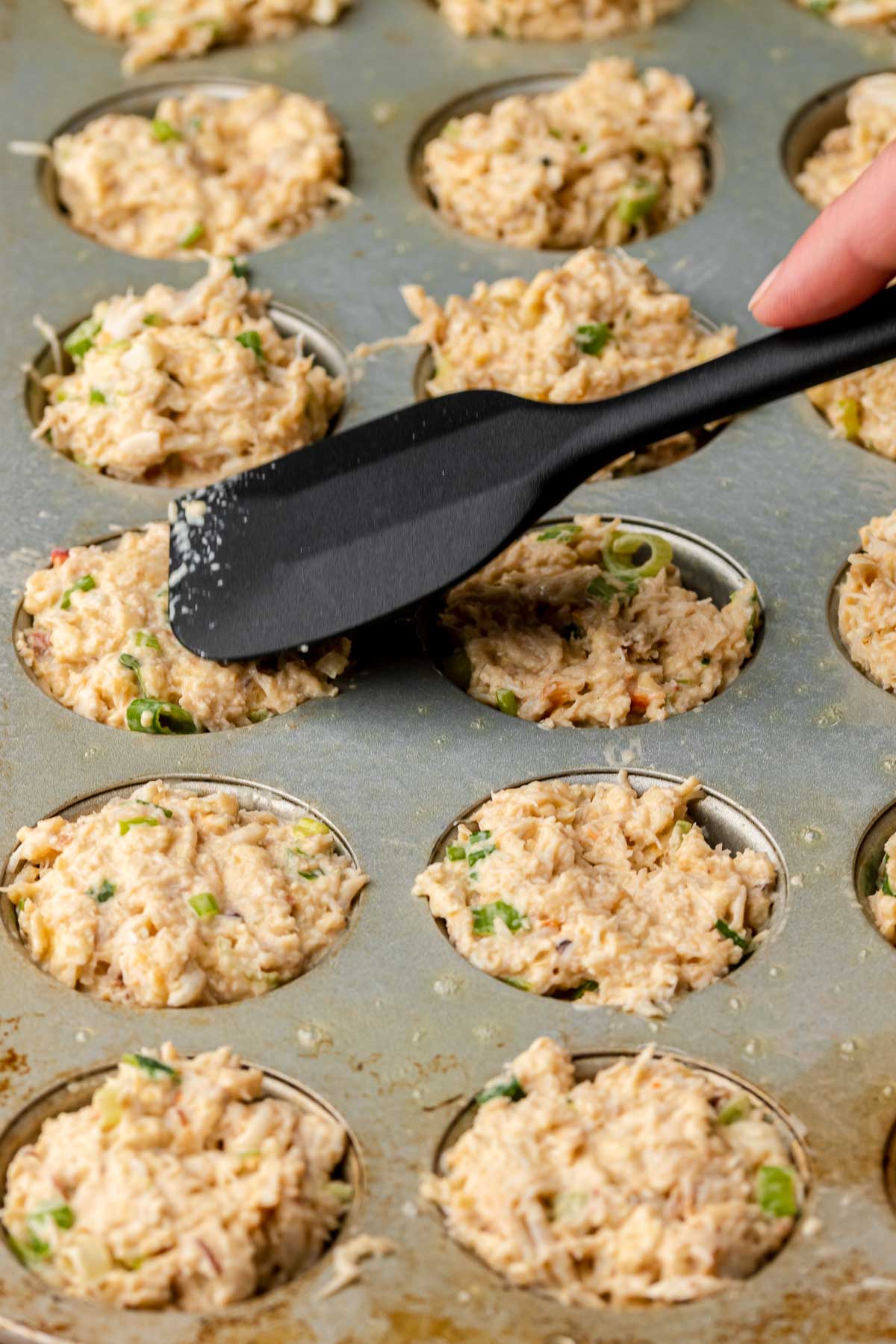 A spatula presses down crab cakes in a muffin tin.
