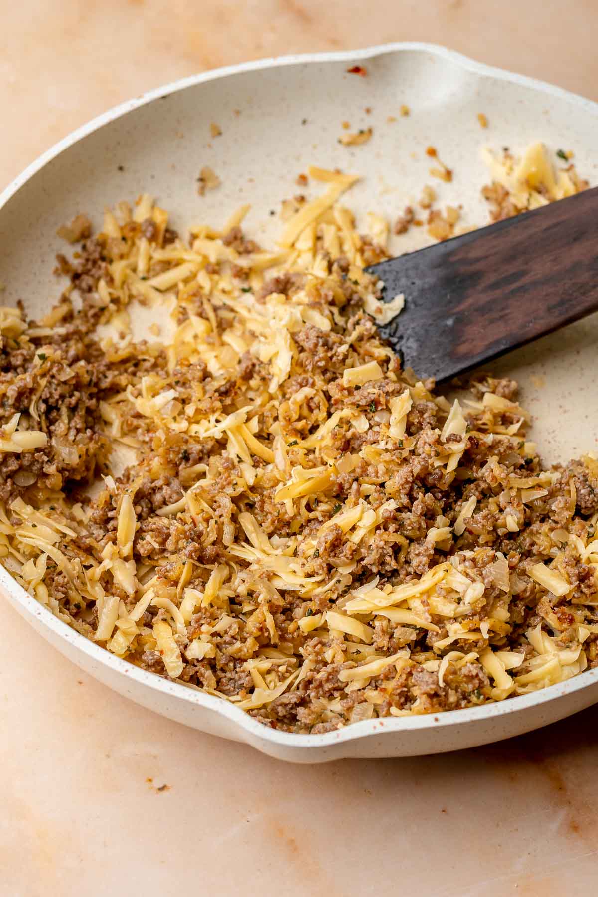 A spatula tosses cheese in sausage filling in a pan.