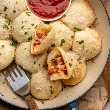 Pizza bites baked in a dish with marinara and a serving utensil. One ball is cut open to show the filling.