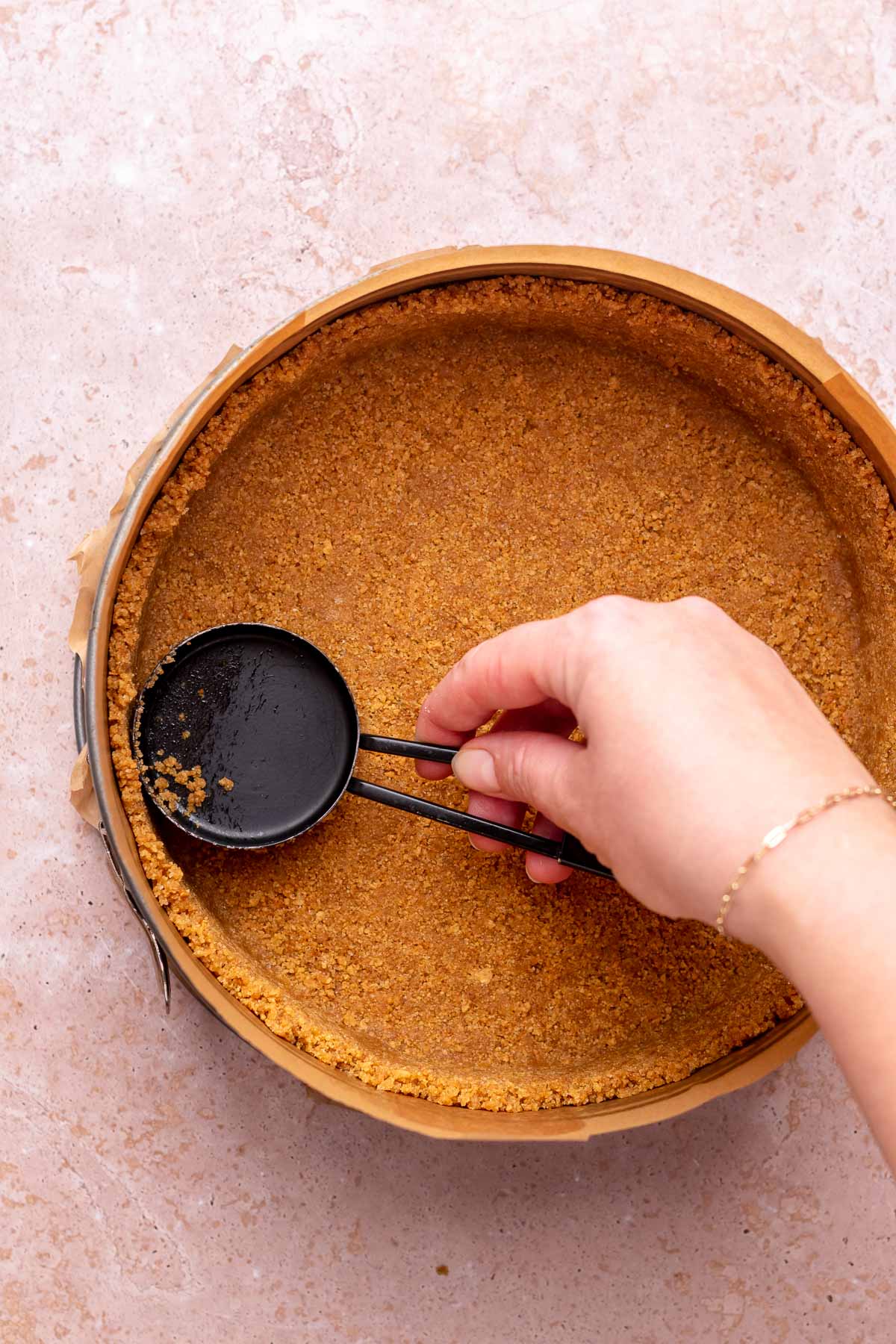 A hand uses a measuring cup to press graham cracker crust into a pan.