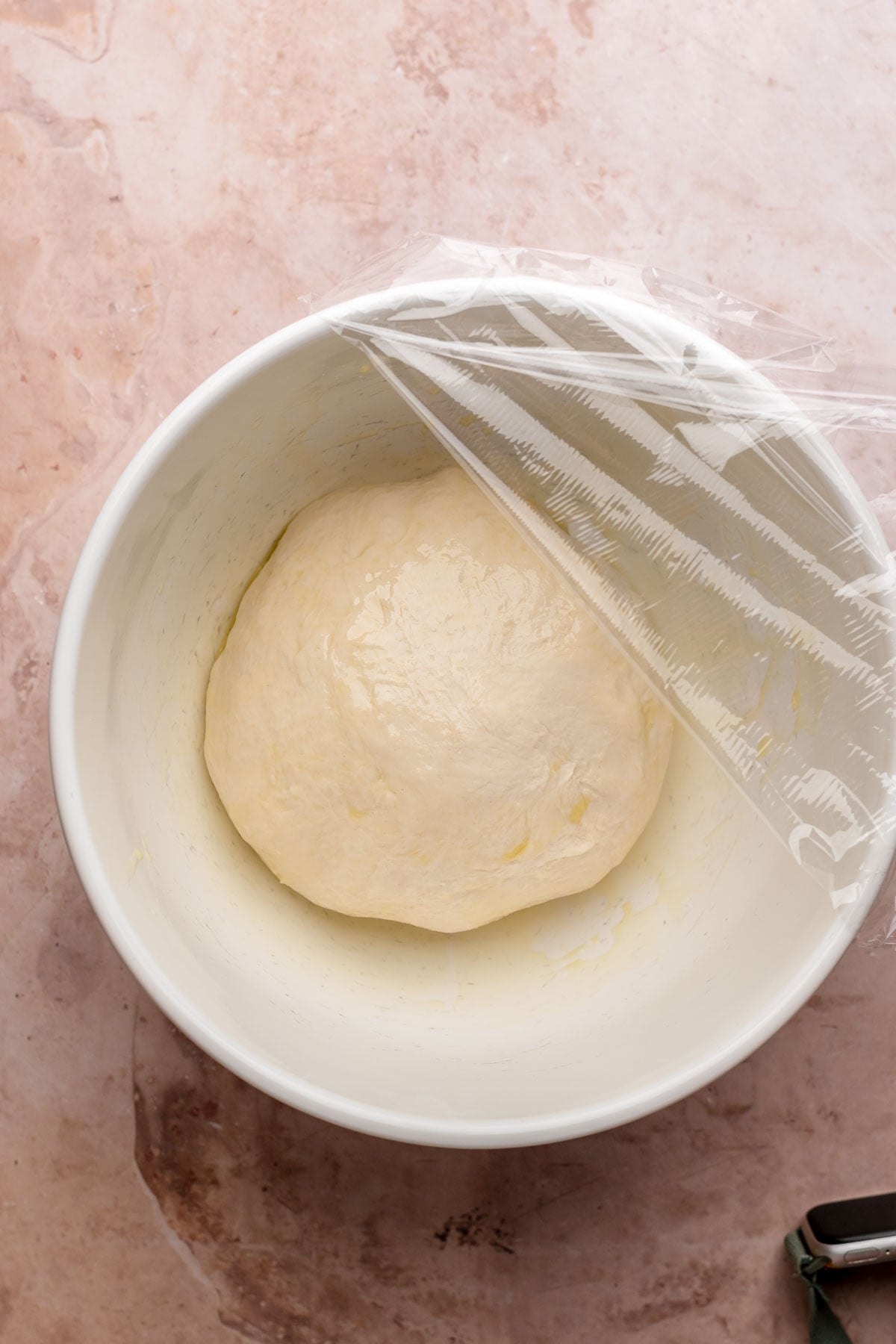 A dough ball in a greased bowl with plastic wrap on top.