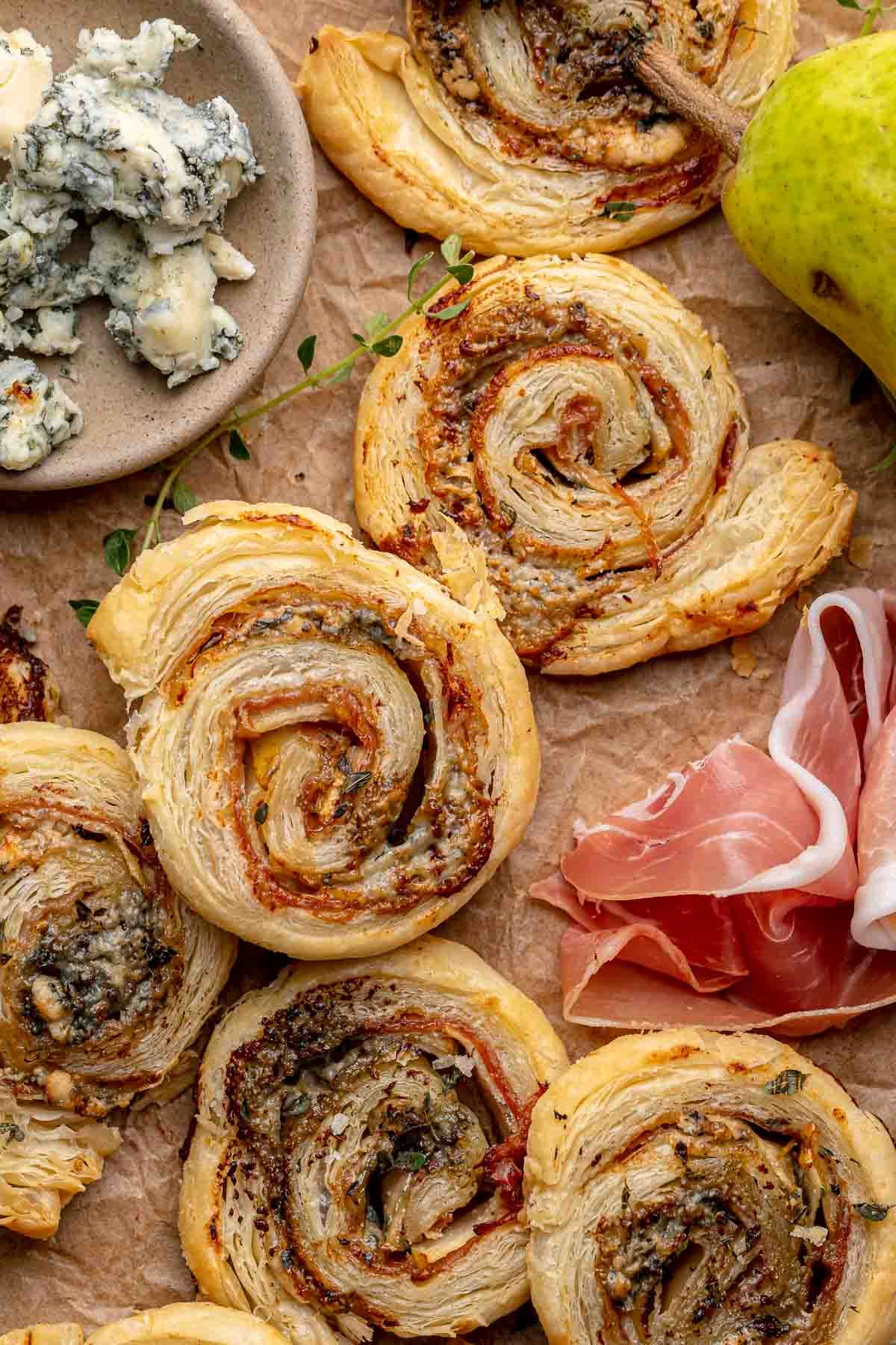 A stack of puff pastry pinwheels on parchment paper. Gorgonzola and prosciutto sit next to it.
